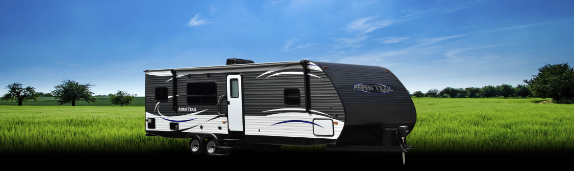 Black & white Aspen Trail trailer parked in a flat, grassy field on a sunny day.